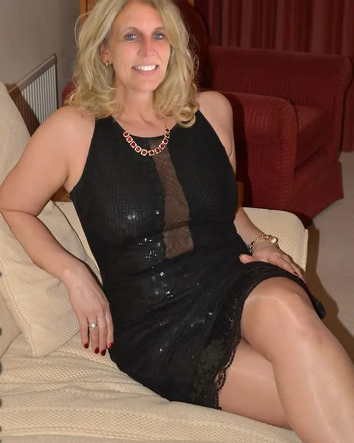 Femme aux cheveux blonds courts, en robe noire à sequins, assise sur un canapé beige, souriant à la caméra.