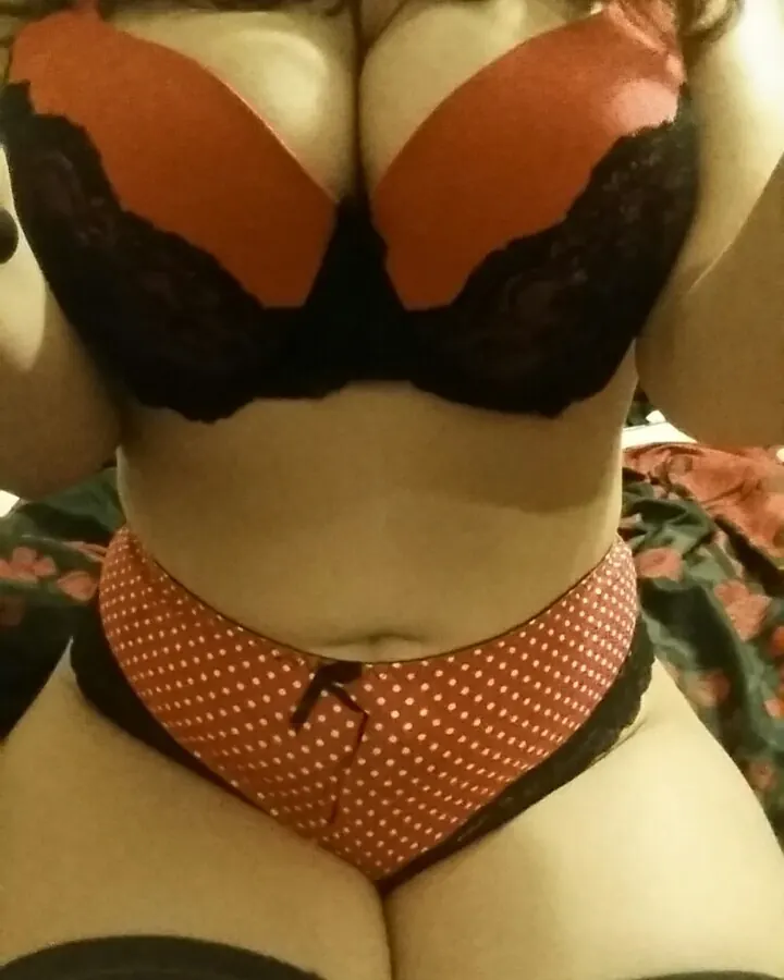 Femme vue de face en lingerie orange et noire, avec des bas noirs, allongée sur un lit.