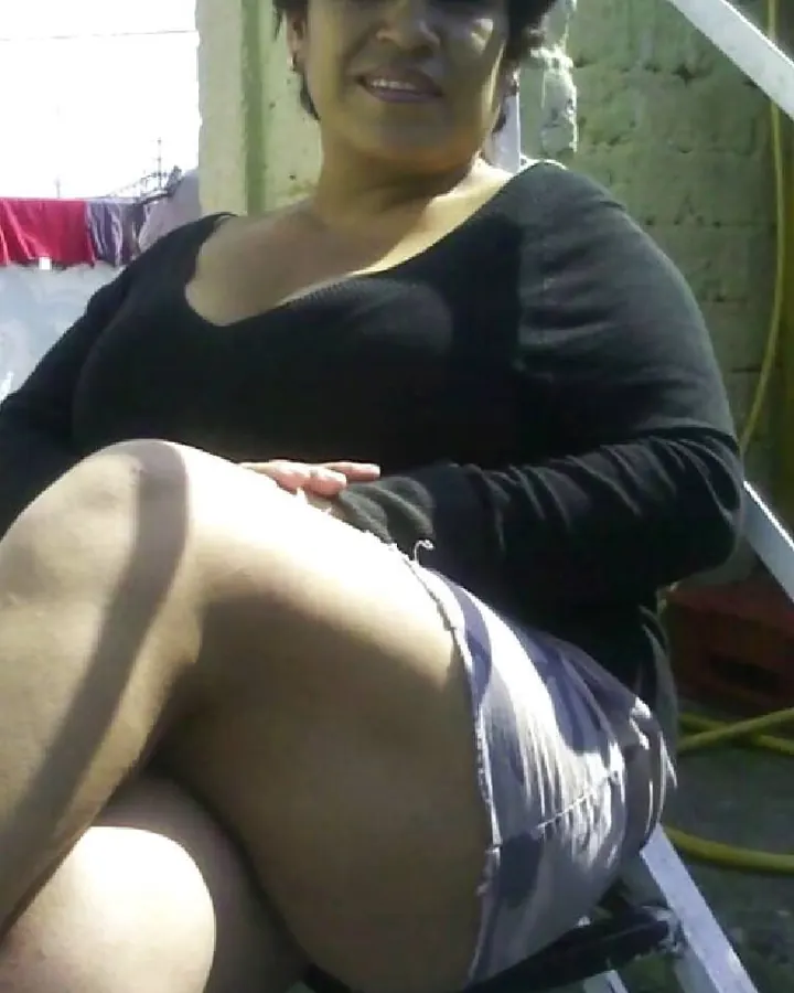 Femme aux cheveux bruns-noirs, souriante, assise sur une chaise, vêtue d'un haut noir, les jambes croisées, dans un jardin, avec un sac à main noir sur les genoux.