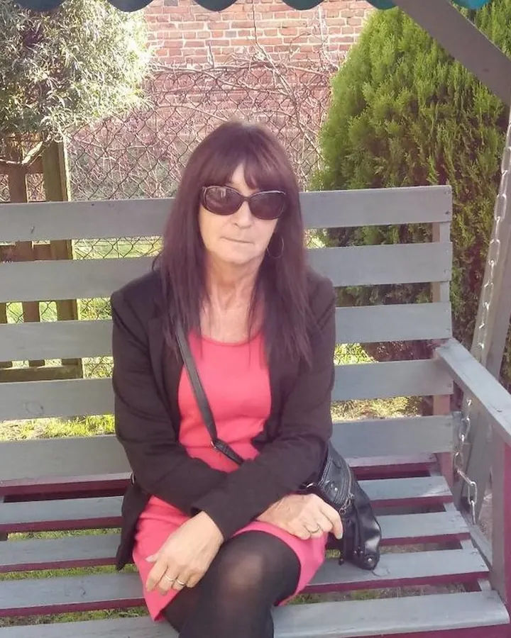 Femme aux cheveux bruns-noirs, portant des lunettes de soleil, assise sur un banc en bois, vêtue d'un haut rose et d'une veste noire, les jambes croisées, dans un jardin.