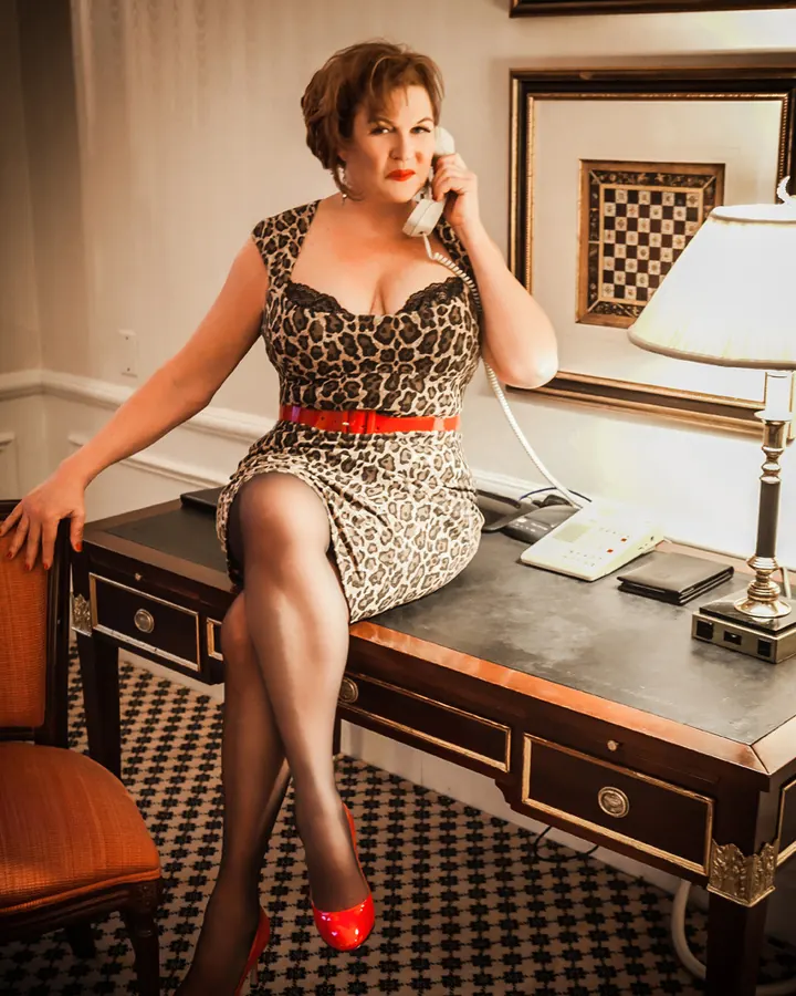 Femme aux cheveux blonds bouclés, en robe léopard, assise sur un bureau en bois, tenant un téléphone blanc à l'oreille.