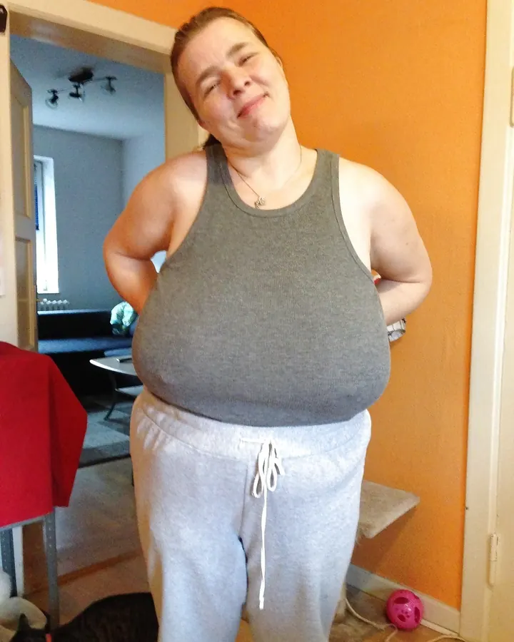 Femme aux cheveux bruns courts, portant un débardeur gris et un pantalon de survêtement gris, debout dans un intérieur avec un mur orange, les mains sur les hanches.