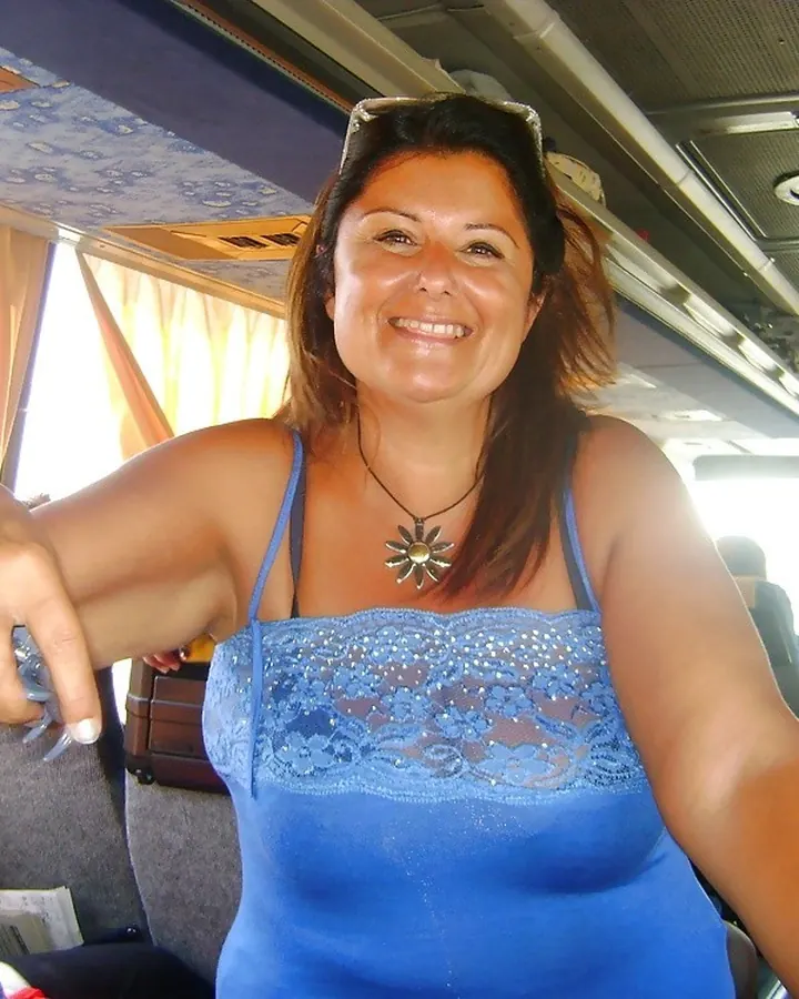 Femme aux cheveux bruns longs, portant un haut bleu à dentelle et un collier en forme de soleil, assise dans un bus avec des lunettes de soleil sur la tête.