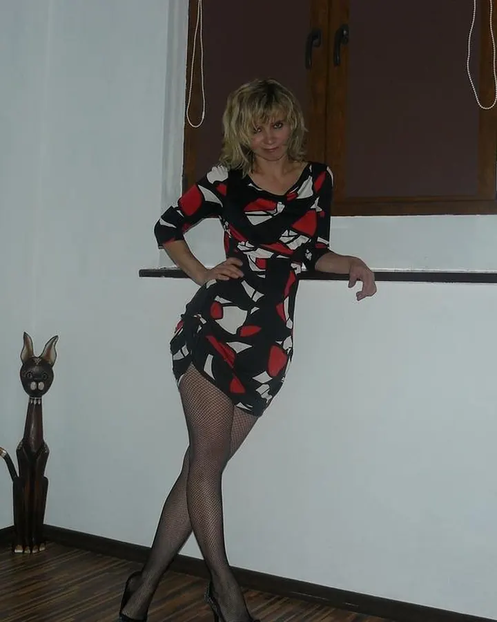 Femme aux cheveux blonds en robe noire, collant noir, chaussures à talons, debout près d'une fenêtre, main sur la hanche, regard vers le côté