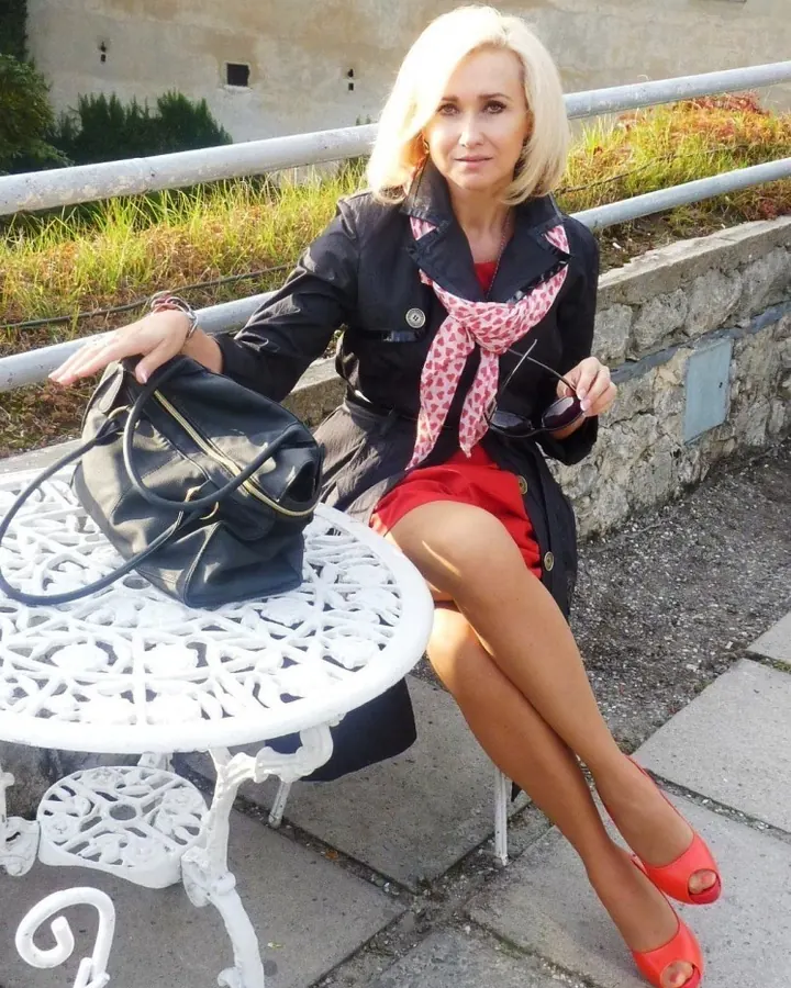 Femme aux cheveux blonds assise sur un banc en bois près d’un lac, vêtue d’une robe noire ajustée, de gants noirs et de talons hauts.