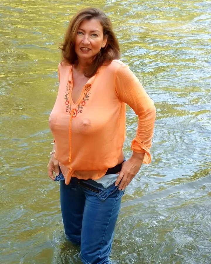Femme aux cheveux blonds assise sur un banc en bois près d’un lac, vêtue d’une robe noire ajustée, de gants noirs et de talons hauts.