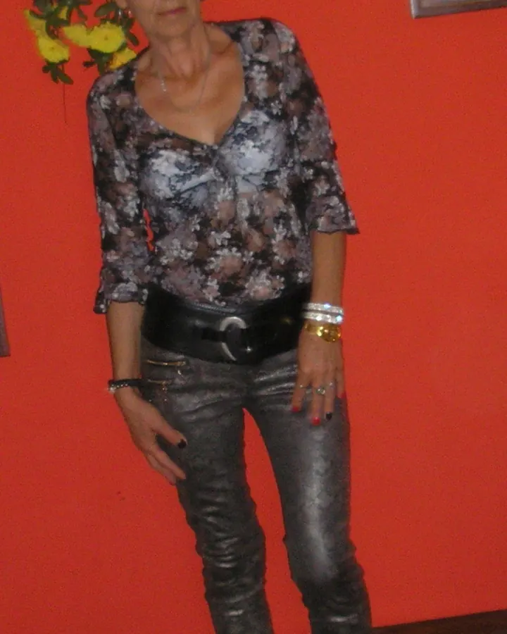 Femme aux cheveux noirs, portant une chemise à motifs et un pantalon en cuir argenté, debout devant un mur rouge.