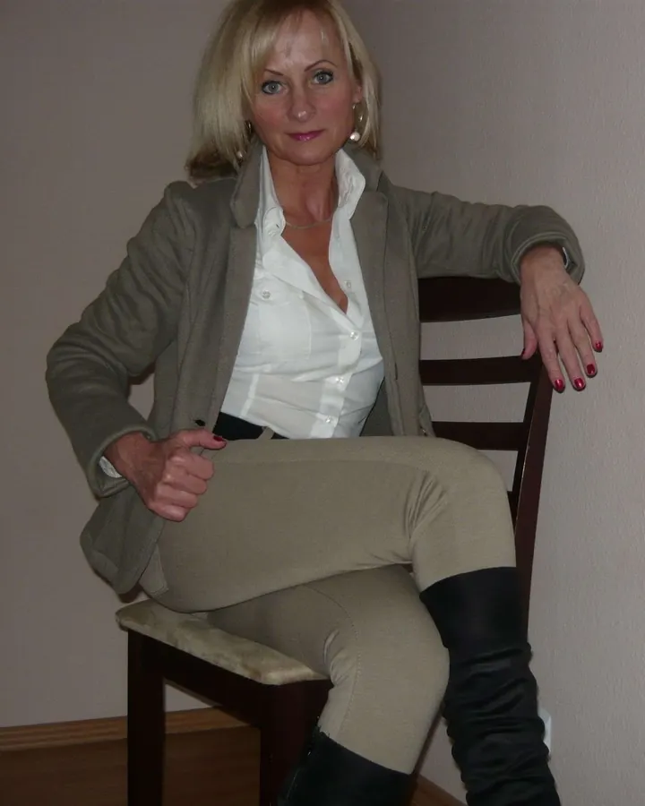 Femme blonde assise sur une chaise, portant un blazer beige et une chemise blanche, les jambes croisées, regardant la caméra.