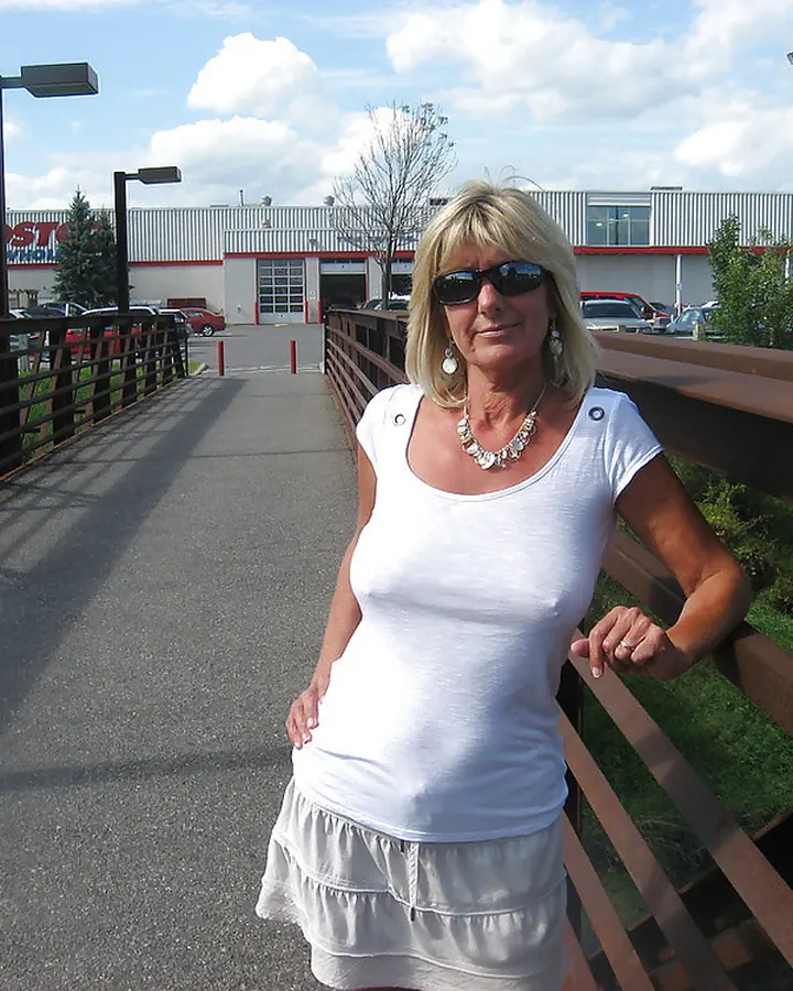 Femme blonde portant un haut blanc, debout sur un pont métallique, regardant la caméra avec un sourire.