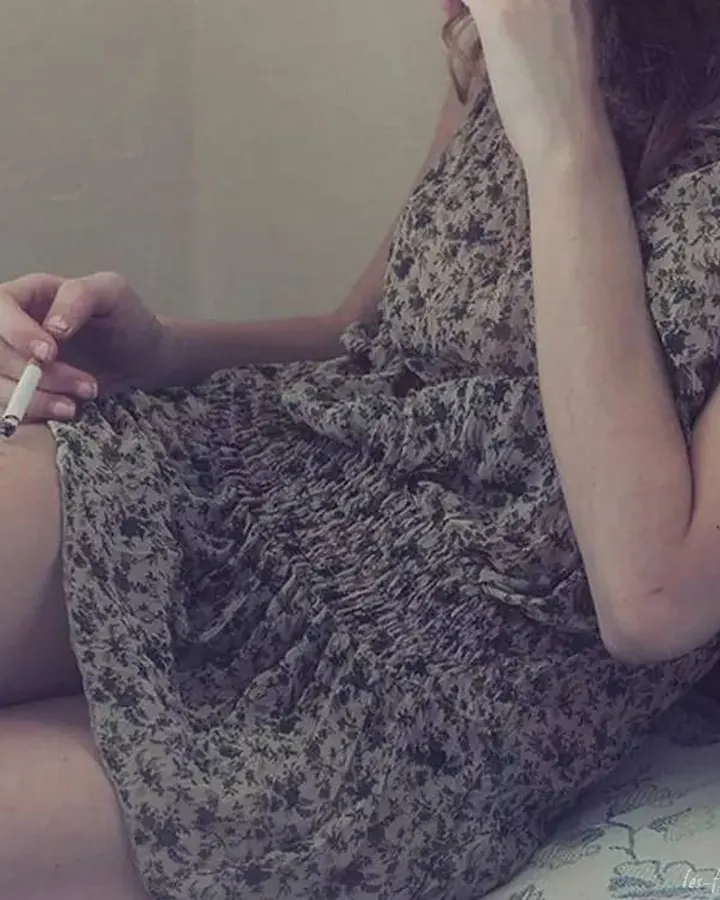 Femme assise, portant une robe à motifs floraux, tenant une cigarette dans sa main droite, sur un canapé.