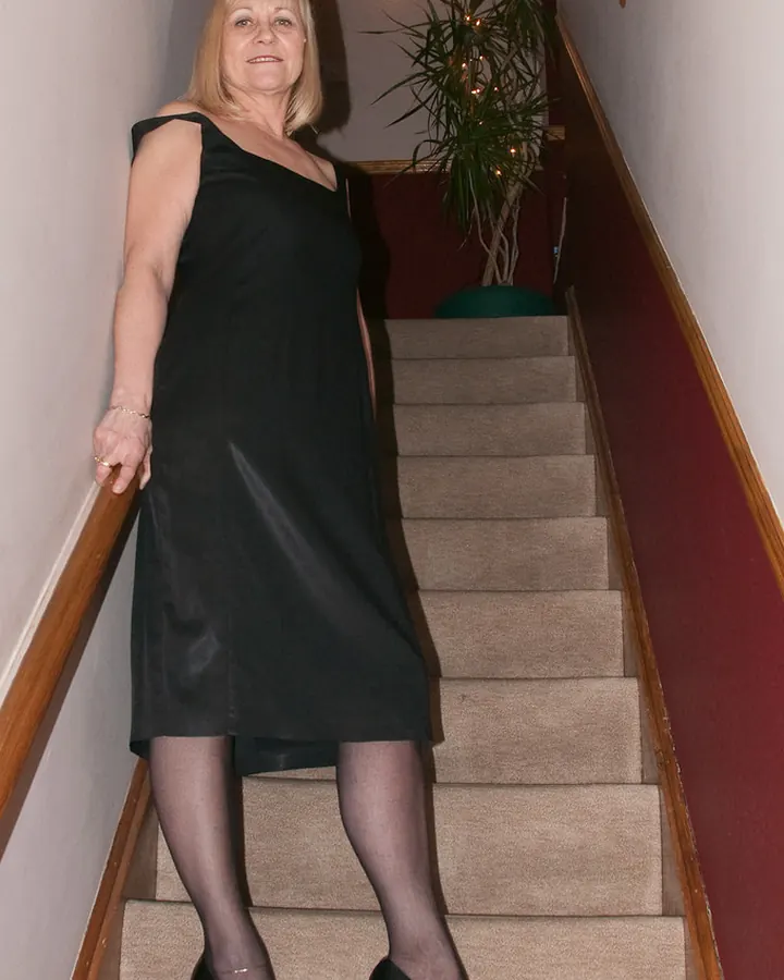 Femme blonde en robe noire, descendant un escalier, souriante, avec des chaussures noires à talons.