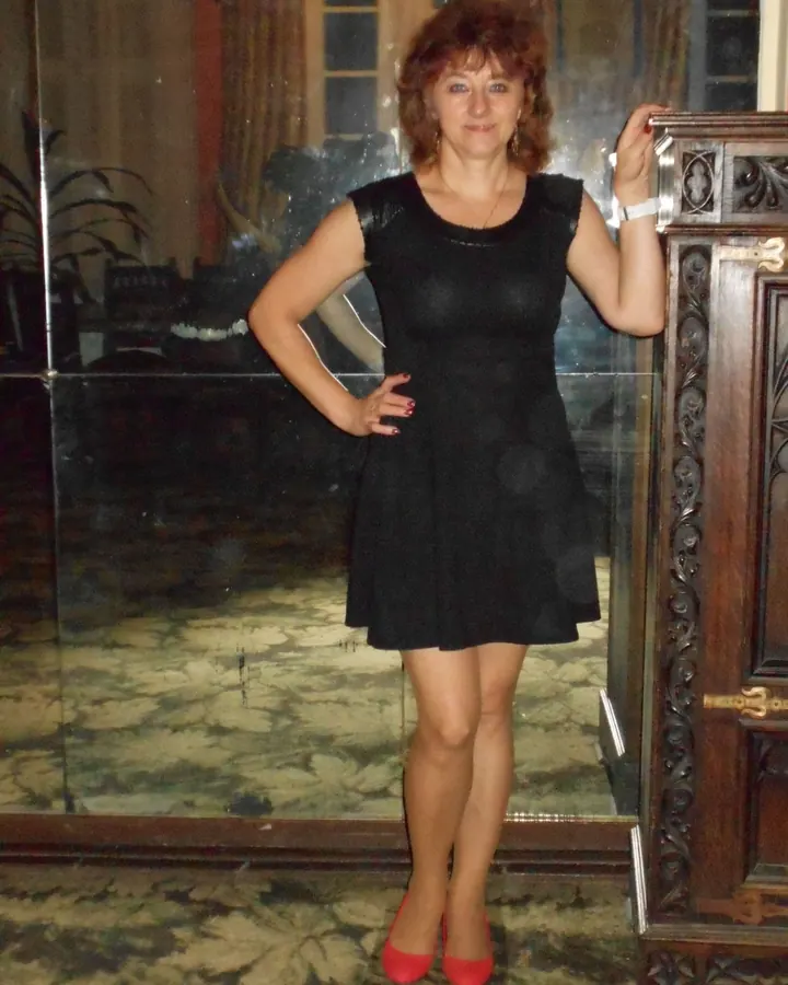 Femme en robe noire satinée, assise sur un canapé, portant des collants noirs et des chaussures rouges.