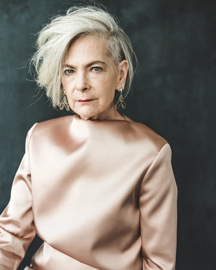 Femme aux cheveux blancs courts, vêtue d'une robe rose pâle, regardant l'objectif avec un regard intense, devant un fond sombre.