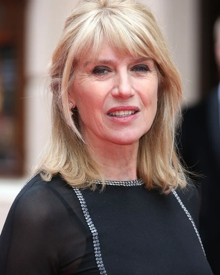 Femme aux cheveux blonds, portant une robe noire transparente avec dentelle, assise sur un canapé noir, regardant l'objectif avec un sourire discret.