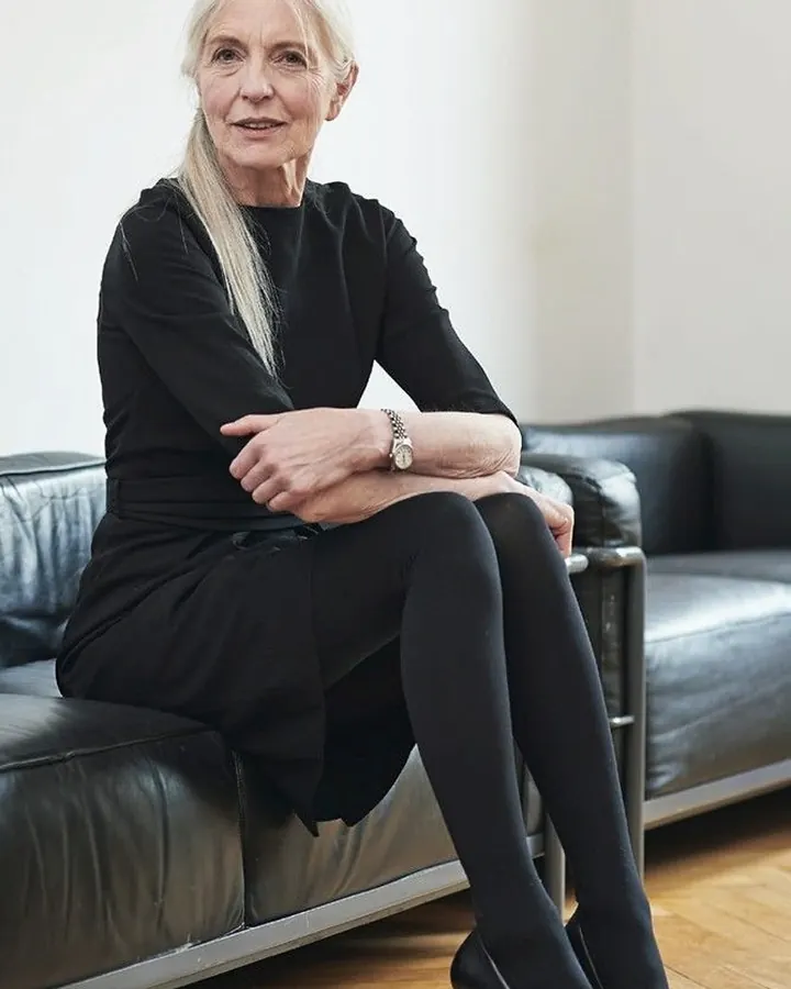 Femme aux cheveux blancs, vêtue d'une robe noire, assise sur un canapé en cuir noir, regardant l'objectif avec un sourire discret.