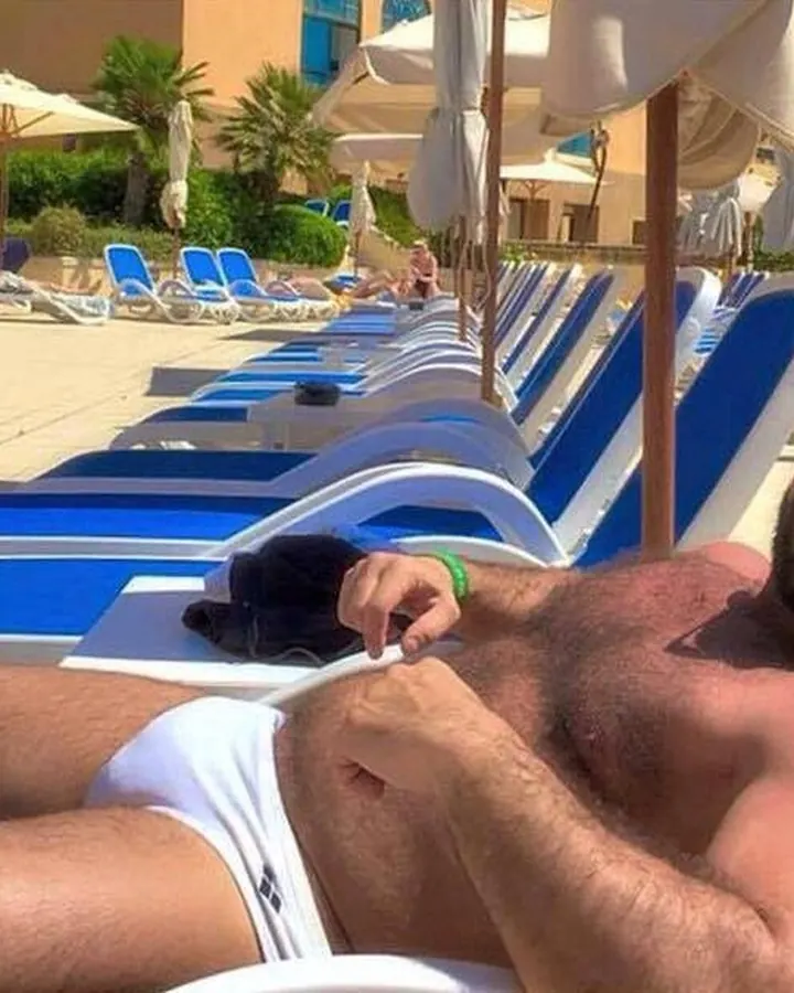Homme torse nu, allongé sur une chaise longue bleue, portant des lunettes de soleil, au bord d'une piscine sous un ciel bleu.