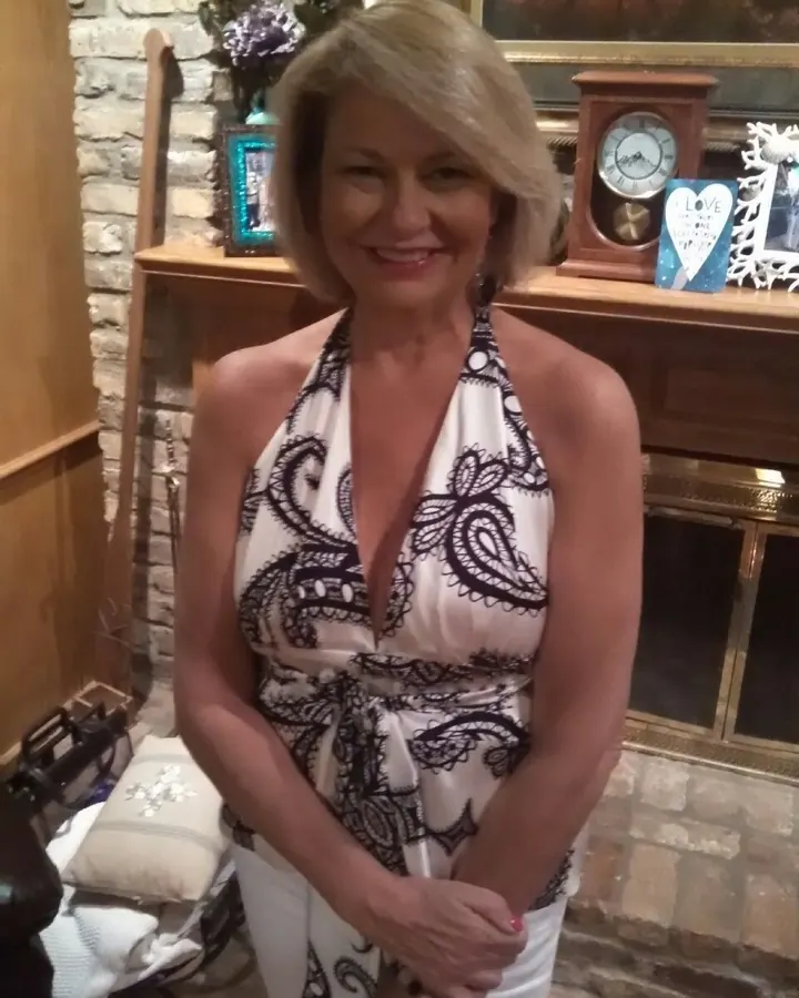 Femme aux cheveux blonds portant une robe bleue, debout près d’un meuble avec une lampe et un téléphone.