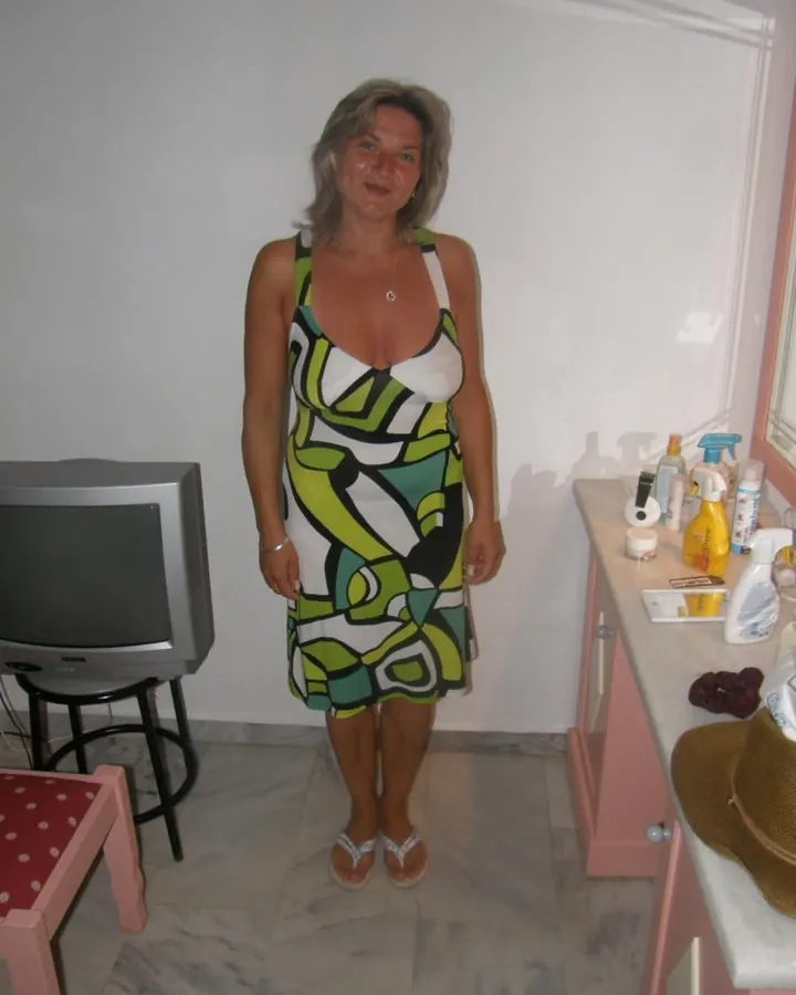 Femme aux cheveux gris clair debout dans une pièce, portant une robe colorée à motifs géométriques, près d’un téléviseur.