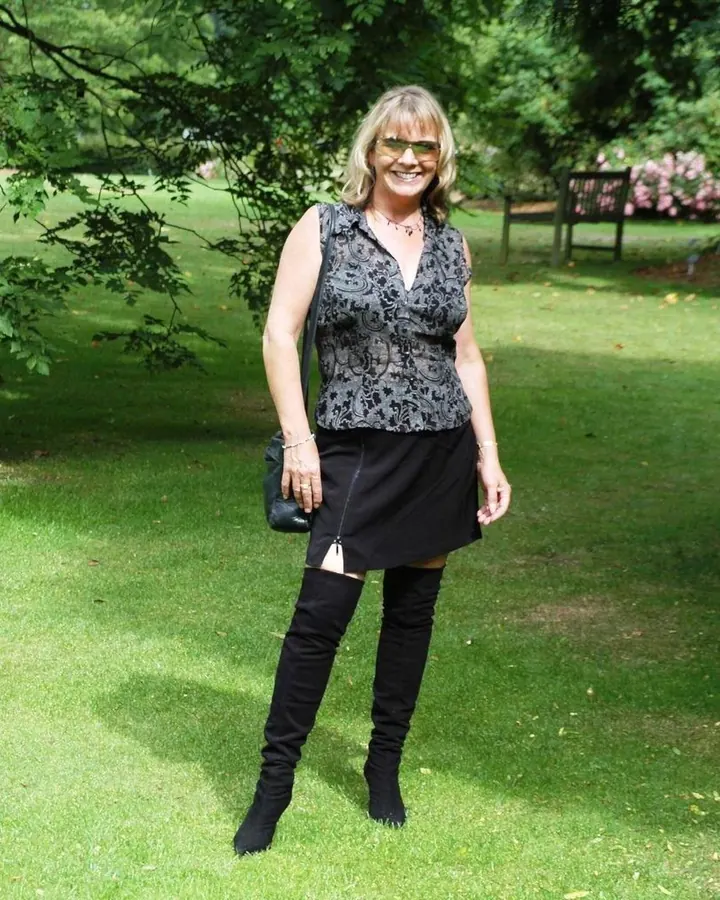 Femme aux cheveux blonds, vêtue d'une tenue noire, posant dans un jardin avec des lunettes de soleil vertes.