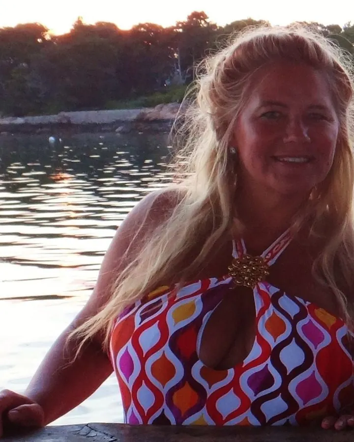 Femme aux cheveux blonds, vêtue d'un maillot de bain coloré, posant au bord d'une eau calme au coucher du soleil.