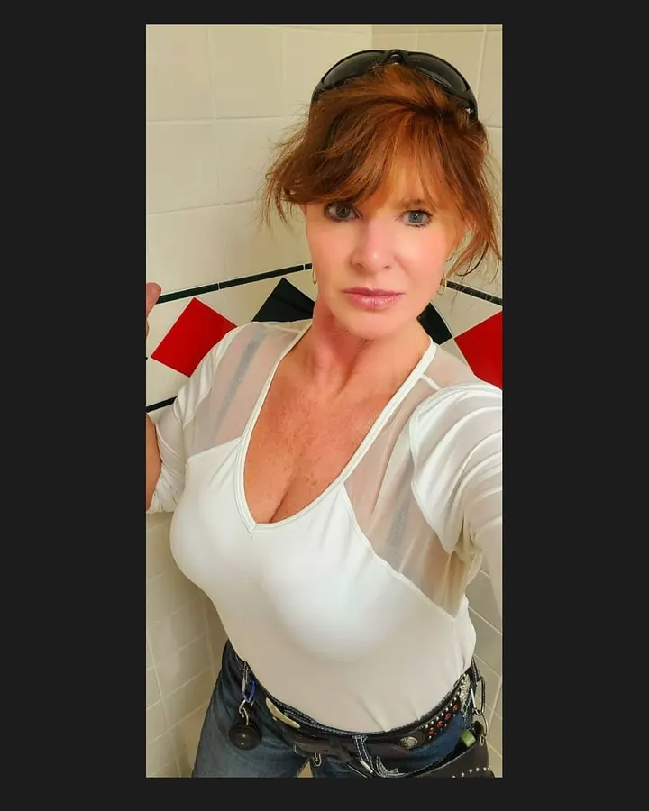 Femme aux cheveux roux, lunettes, en robe rouge satinée, en position accroupie sur un tapis à motifs géométriques.