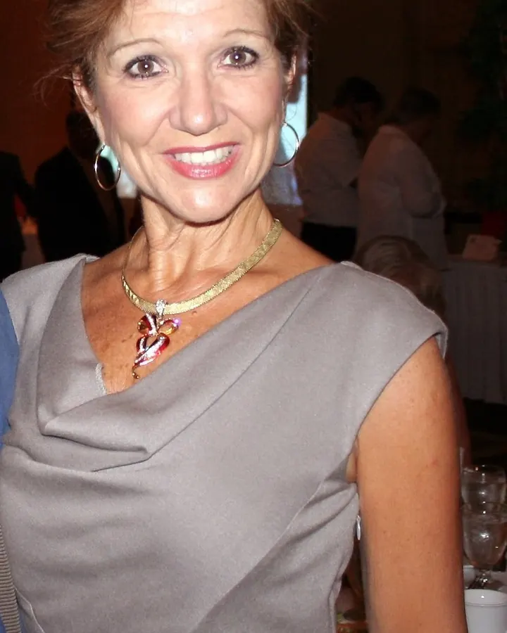 Femme en robe grise, souriante, avec un collier coloré, dans une salle de réception.